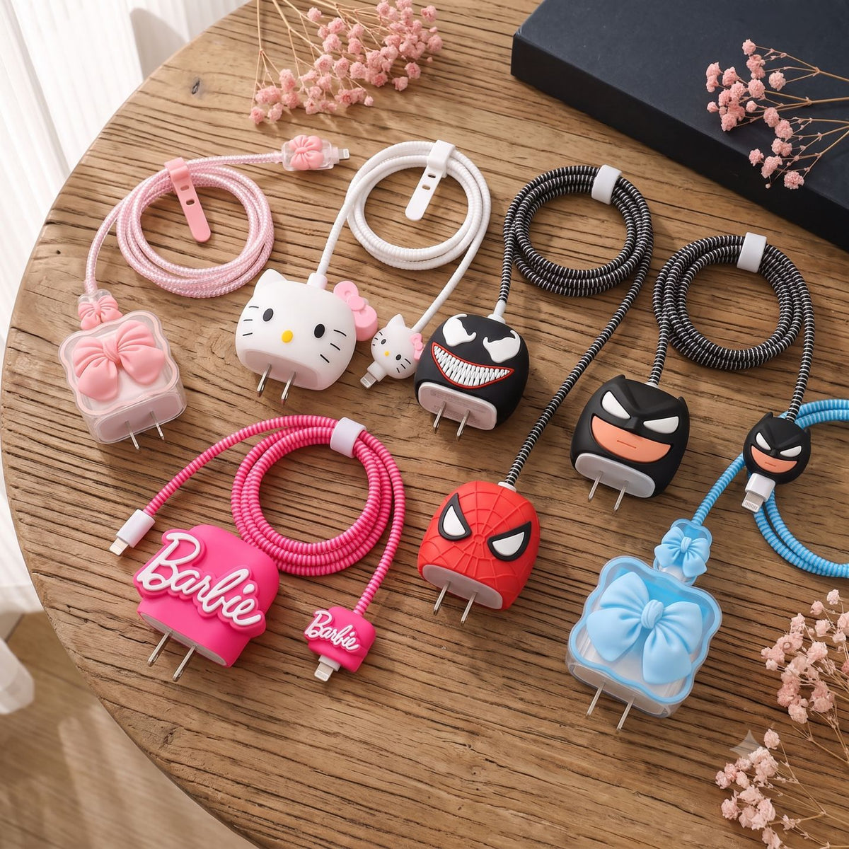 Six colorful character-plug extension cords arranged on a wooden tabletop. The cords feature cartoon faces and bows: Hello Kitty, Venom, Batman, Spider-Man, Barbie, and a blue bow-tie Batman design.