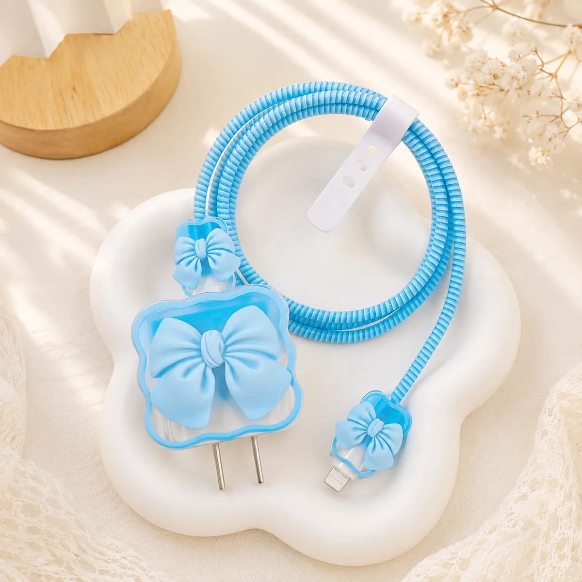 A light blue silicone cable protector with a bow design, shown with a coiled blue cable and a USB-C connector, displayed on a white cloud-shaped tray.