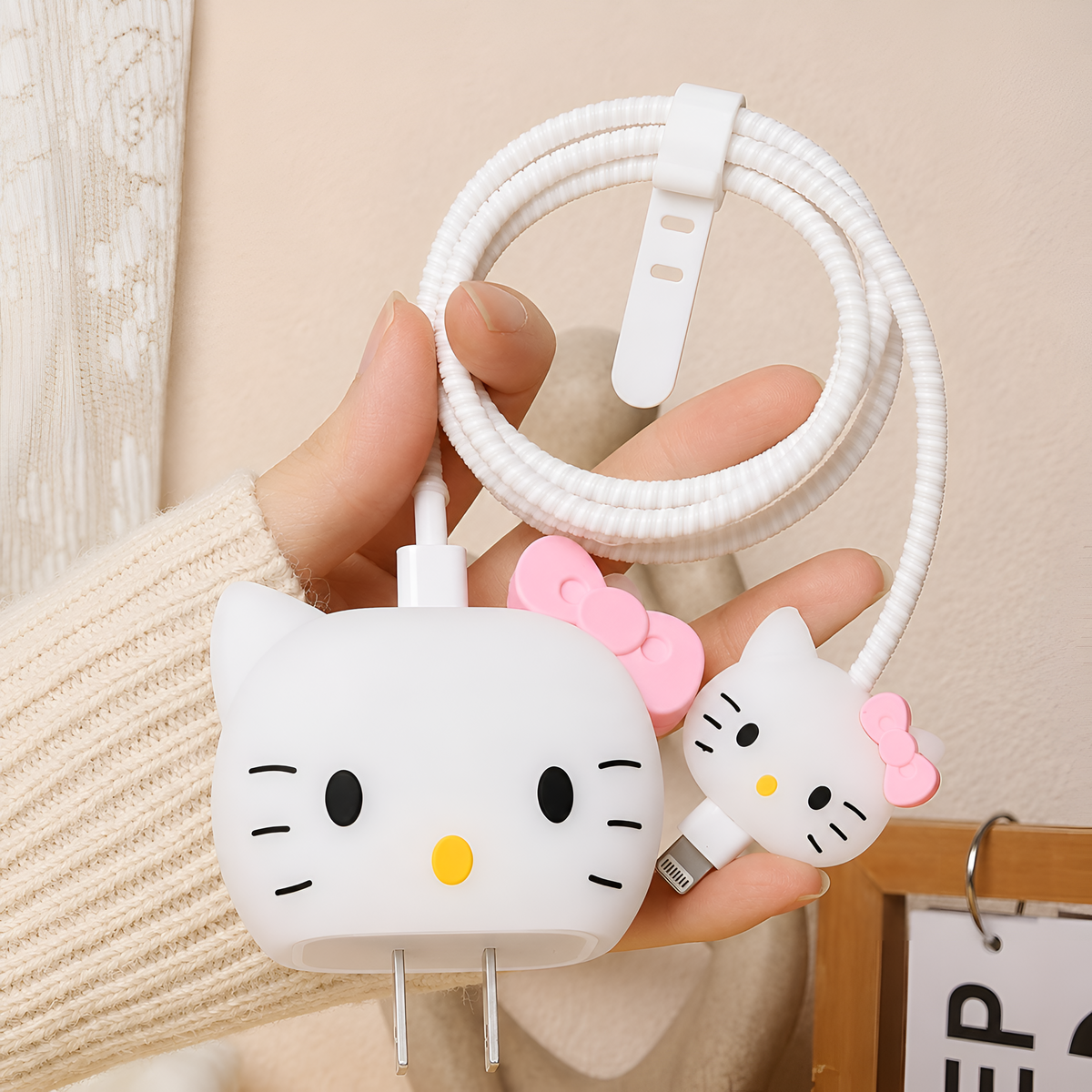 A hand holding a white coiled charging cable with two Hello Kitty-shaped USB-C adapters, one with a pink bow and one with a black bow, against a soft beige indoor background.