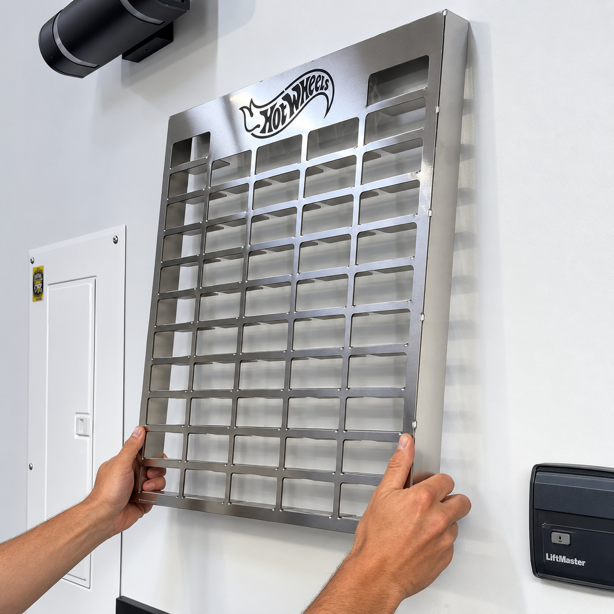 A person's hands are holding a large stainless steel wall-mounted organizer rack with a 5x5 grid of small compartments. The rack features the Hot Wheels logo at the top and is mounted on a white wall in an indoor setting.
