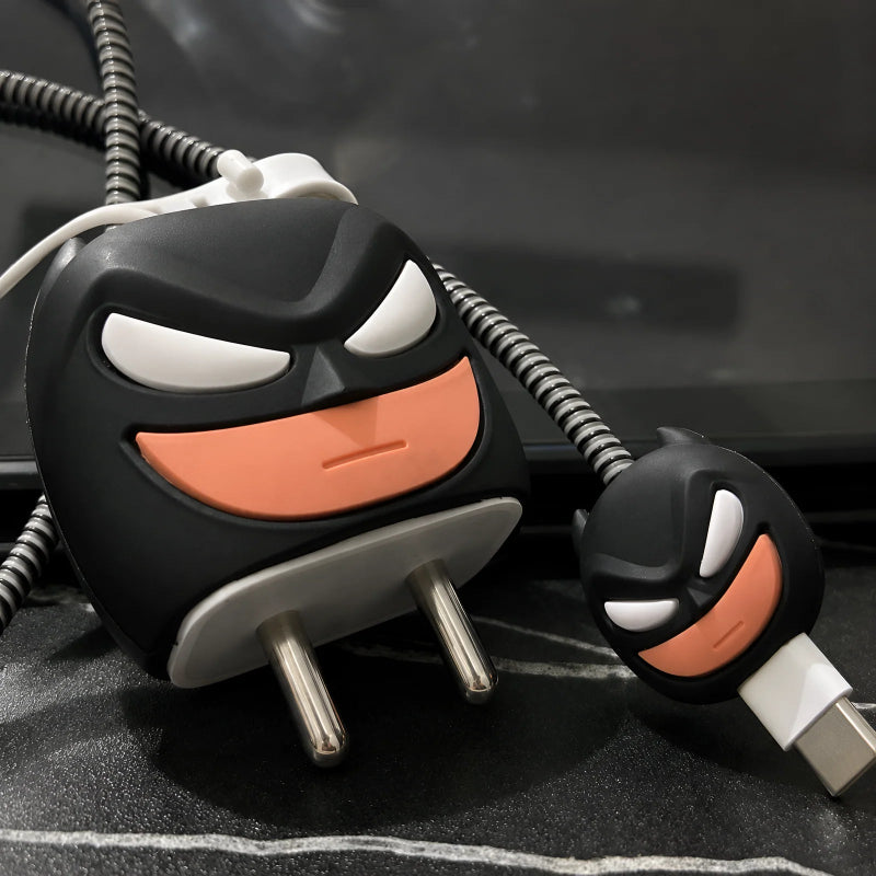 A close-up of a black and white Batman-themed USB charger with a matching USB-C cable. The charger features a black face with white eyes and a wide orange smile, and the cable has a matching connector with the same design.