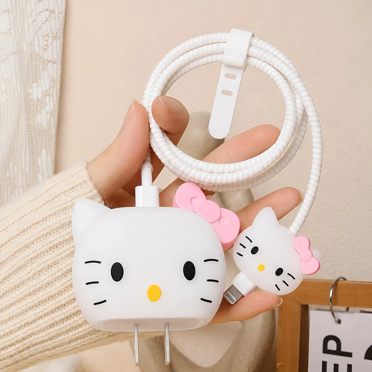 A hand holding a white coiled charging cable with two Hello Kitty-shaped USB-C adapters, one with a pink bow and one with a black bow, against a soft beige indoor background.