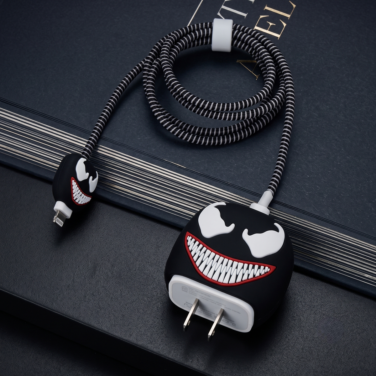 A coiled black and white braided charging cable with a Venom-inspired character face design on the plug and connector ends, photographed on a dark textured surface.