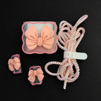 A hair accessory set featuring a large peach-pink bow, two smaller matching bows in clear clips, and a coiled beaded elastic cord with a white plastic connector, all arranged on a solid black background.