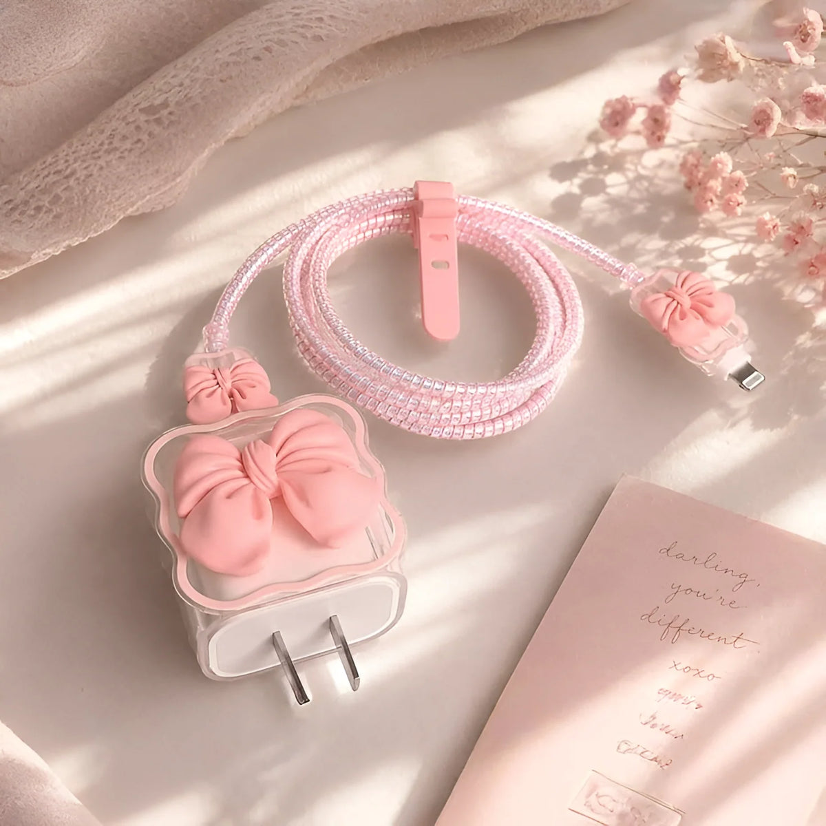 A pink decorative charging cable with a large bow-shaped plug and matching bow details, arranged on a soft beige surface with lace fabric, dried flowers, and a pink card in the background.