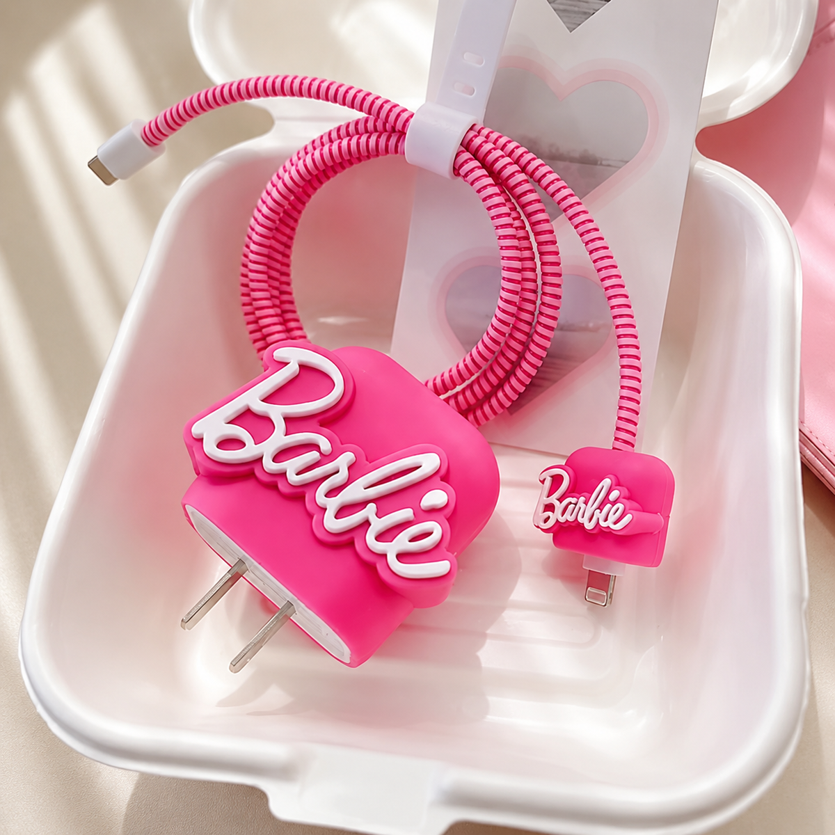 A pink coiled charging cable with a matching plug, both featuring a 3D Barbie logo, displayed in a white plastic container.