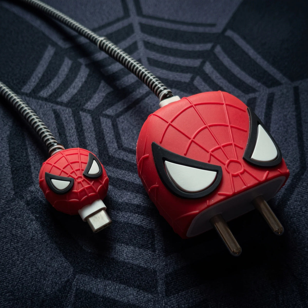A close-up of a red and black Spider-Man-themed charging cable with a USB-C connector, featuring a large red plug shaped like Spider-Man's face on a dark patterned fabric background.