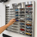 A stainless steel Hot Wheels wall-mounted display board with multiple recessed slots holding assorted toy cars, shown with a person's hand reaching for one of the toy cars.