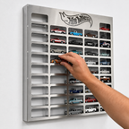 A silver metal Hot Wheels model car display wall mounted on a white wall, with a hand holding a small toy car in front of several compartments. The wall-mounted rack features the Hot Wheels logo at the top and holds multiple rows of miniature die-cast cars.