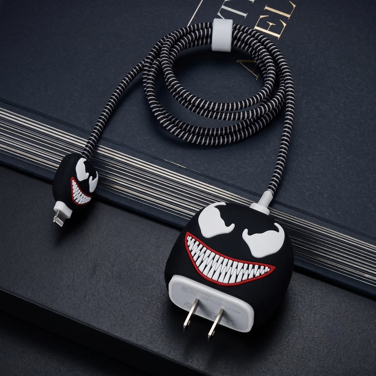 A coiled black and white braided charging cable with a Venom-inspired character face design on the plug and connector ends, photographed on a dark textured surface.